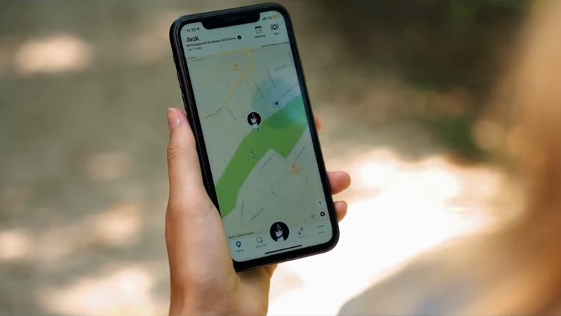 A person holds a smartphone showing a real-time dog tracking app with location updates on a forest path